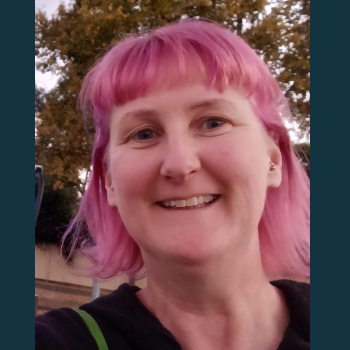 Smiling person with pink hair outdoors, wearing a black top with trees in background.